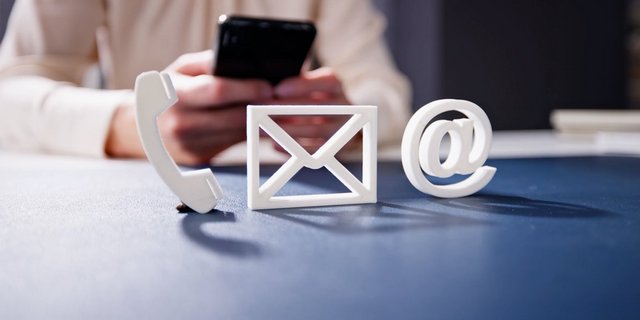 Eine Hand hält ein Smartphone, während daneben Symbole für Telefon, E-Mail und @-Zeichen auf einem Tisch liegen.