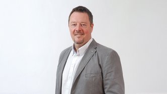 Ein Mann mit einem freundlichen Lächeln trägt einen grauen Blazer und steht vor einem hellen Hintergrund.