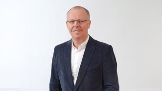 Ein Mann in einem blauen Blazer lächelt freundlich vor einem hellen Hintergrund, was eine positive Ausstrahlung vermittelt.