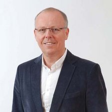Professioneller Mann in einem eleganten Anzug lächelt freundlich Ein lächelnder Mann in einem blauen Blazer steht vor einem hellen Hintergrund und strahlt Selbstbewusstsein aus.