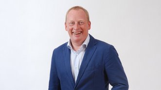 Ein lächelnder Mann in einem blauen Anzug steht vor einem hellen Hintergrund und strahlt Selbstbewusstsein aus.